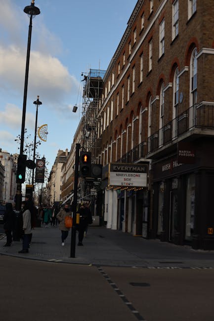 A wet street scene in Marylebone, London, showing a curved road lined with black bollards along the pavement. To the left, a small park area with leafless trees and green shrubbery surrounds a statue.