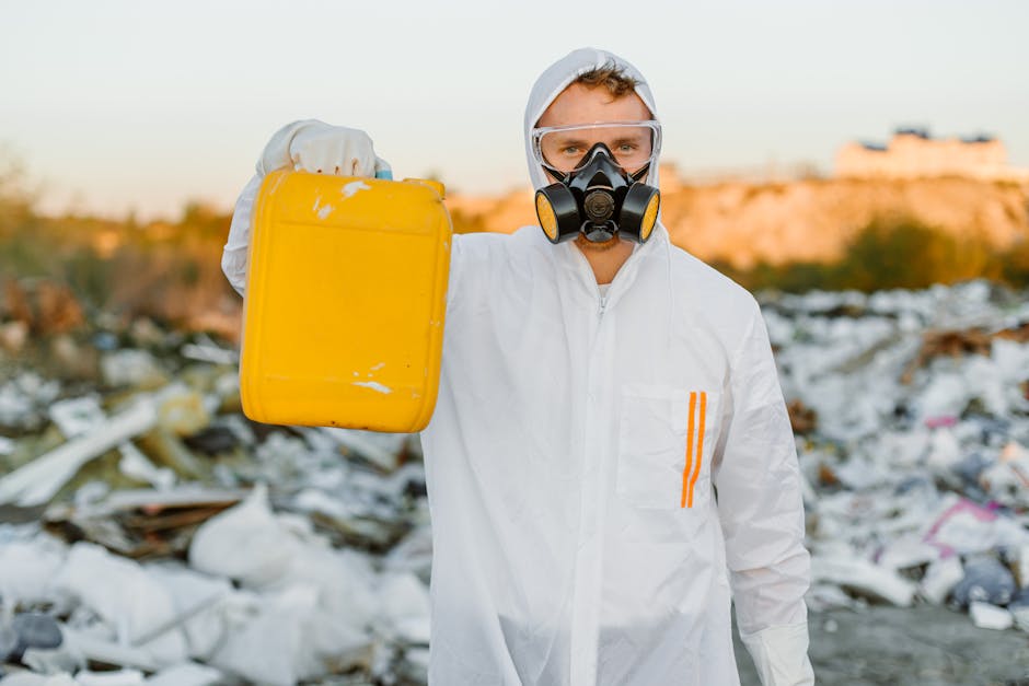 A person wearing a white hazmat suit, protective goggles, and a respirator mask is standing amid a large area of scattered waste and debris, which includes plastic bags, cardboard boxes, and various d