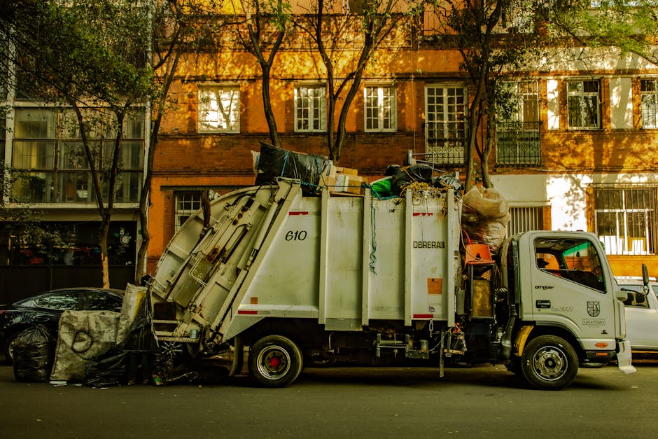 A blue recycling bin positioned outside a white moving van, which is parked on a street pavement, during a home relocation process. The van has a closed rear door, and part of its side panel is visibl