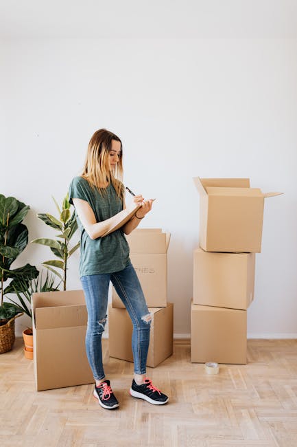 A young woman with shoulder-length blonde hair, wearing a casual green T-shirt, ripped blue jeans, and black sneakers with pink accents, stands inside a room on a light wooden floor. She is writing on a clipboard with a pen, surrounded by stacked cardboard moving boxes of various sizes, some open and others closed. A large potted plant with broad green leaves is positioned to her left in the background. The scene depicts a packing or home relocation process, supported by the presence of packing materials and the setting typical for house removals, as highlighted by the services of Self Storage Marylebone.